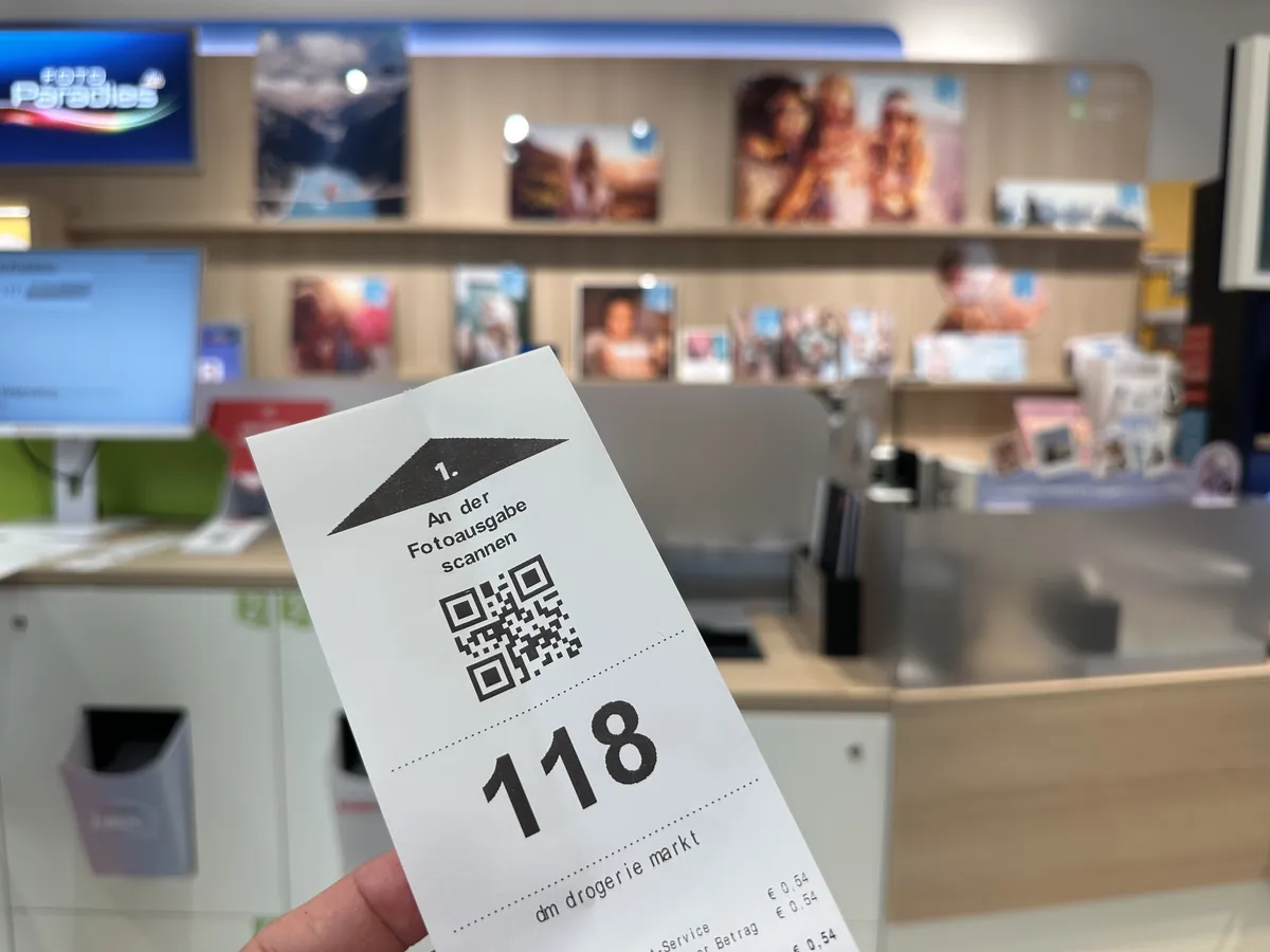 dm drogerie markt Abholticket Nr. 118 mit QR-Code und Hinweis: An der Fotoausgabe scannen. Im Hintergrund das Foto Paradies mit Ausgabefächern.