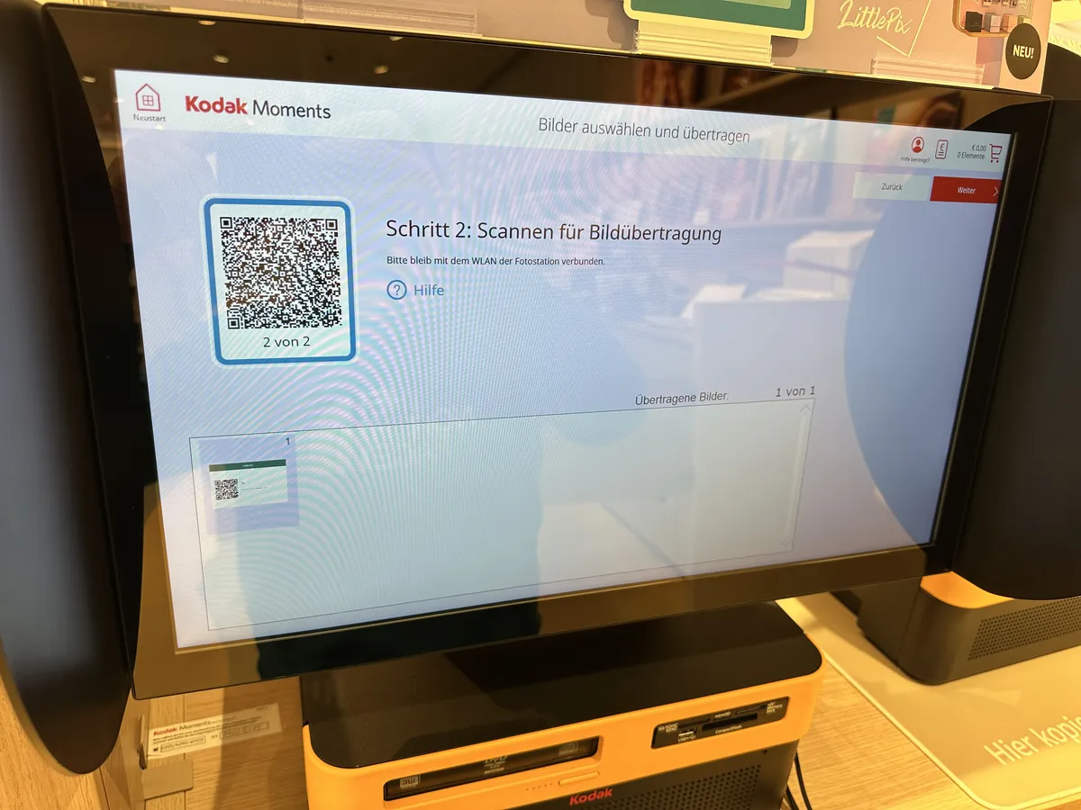 Kodak Moments Fotostation bei dm: Schritt 2 — Scannen für Bildübertragung. Der Bildschirm zeigt einen QR-Code zum Verbinden mit dem Station-WLAN.